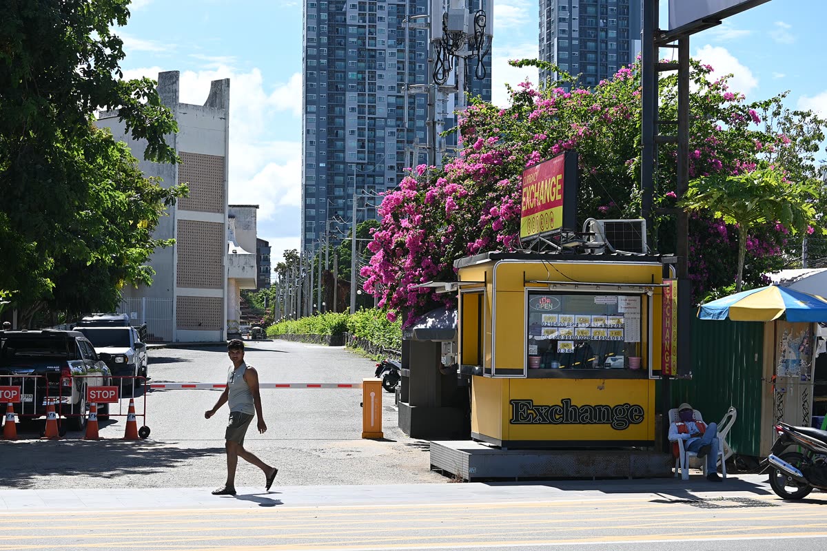 DFC 1464 A pedestrian crosses the street past a bright yellow currency exchange booth with flowering trees and high-rise buildings in the background on a sunny day — Chinese yuan to korean won at 207: what it means for your trip money