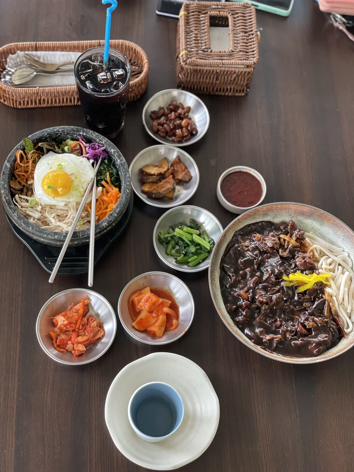 Top-down view of a wooden table with Korean dishes, including bibimbap, jajangmyeon, kimchi side dishes, and an iced drink with a straw. — How much sodium is in kimchi?