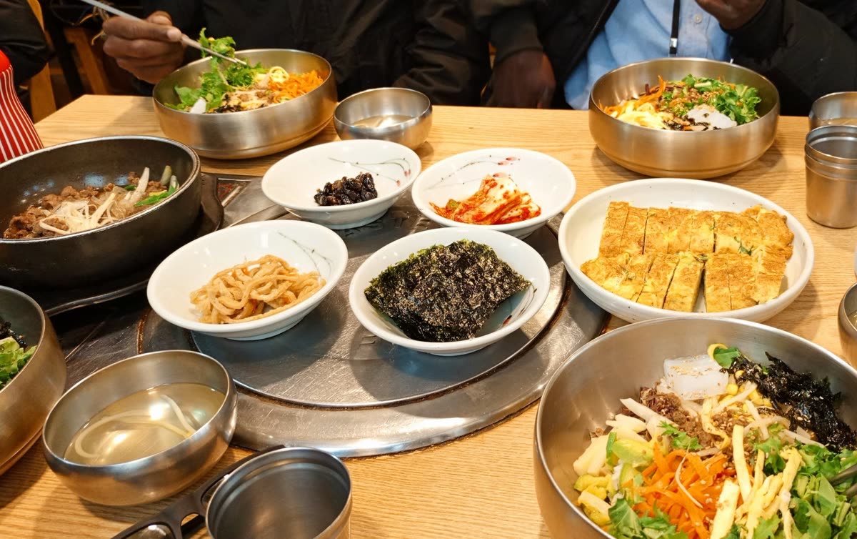 A top-down view of a wooden table filled with various Korean dishes. Large brass bowls contain bibimbap with fresh vegetables and rice, while smaller side dishes of kimchi, seaweed, rolled omelets, and marinated beef are arranged in the center. — Is Korean Food Salty? The One-Line Answer