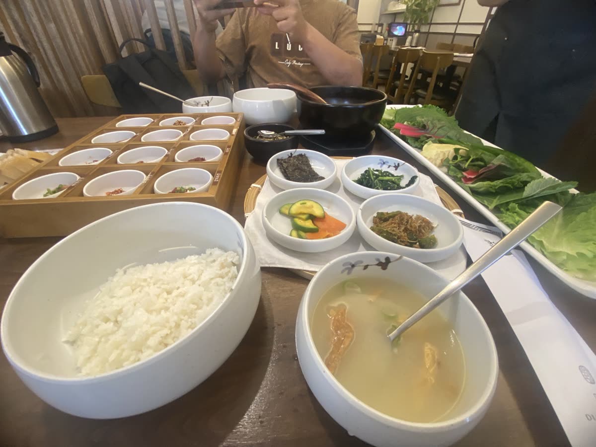 A table setting featuring a variety of dishes: a bowl of white rice, a bowl of light soup with green onions and shredded ingredients, and several small bowls containing side dishes including vegetables and sauces. In the background, there is a wooden tray with additional small bowls, likely for condiments. — Is Korean food good for weight loss? The 5.9% answer on rice and timing
