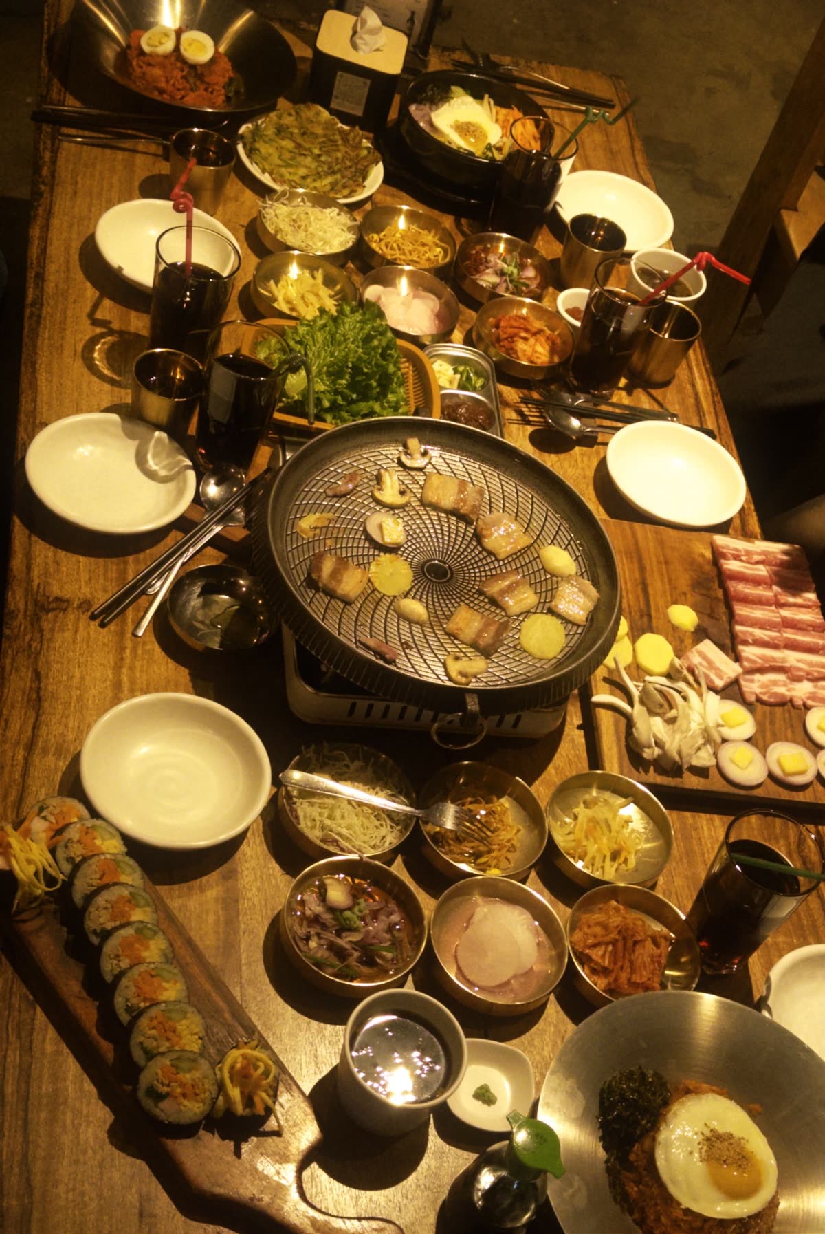 An overhead view of a full Korean BBQ spread on a wooden table featuring a round charcoal grill with meat and mushrooms cooking, raw pork belly slices, multiple banchan side dishes, kimbap rolls, bibimbap and dark drinks. — What Ingredients Do You Need for Korean BBQ — The Full Checklist