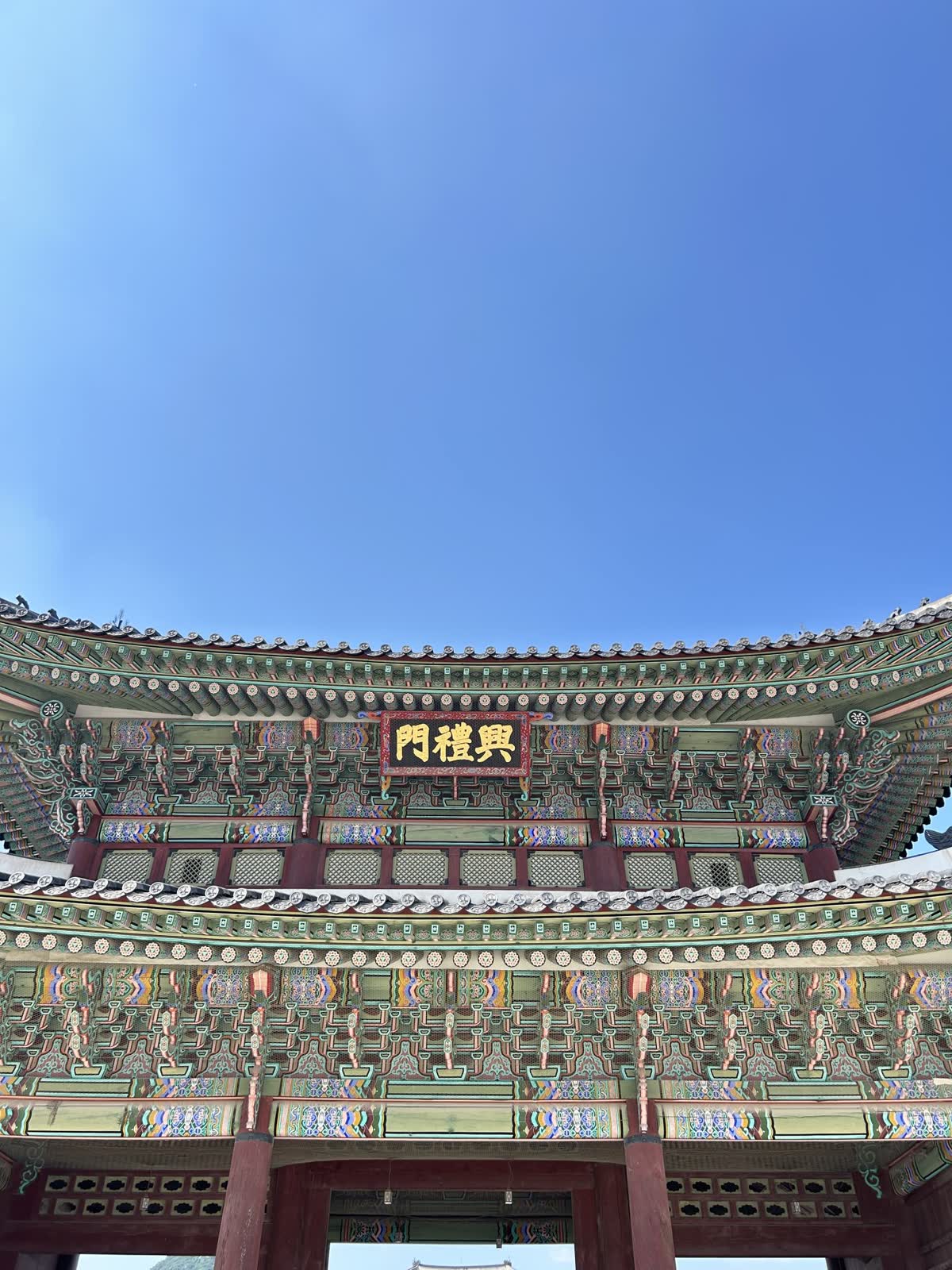 Ornate traditional Korean palace gate under a clear blue sky, featuring intricate green and red designs with a sign displaying Korean characters. — When and where to watch