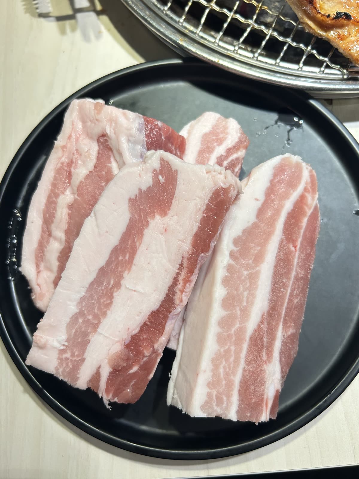 A black circular plate holds four thick pieces of raw pork belly. The pork belly has a distinct layering of pink meat and white fat, showcasing its marbled appearance. The slices are stacked unevenly, with some pieces leaning against each other. — What to Buy When There Is No Korean Butcher